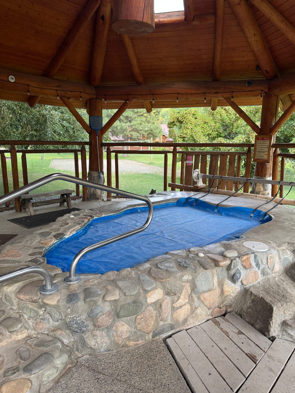 Covered outdoor soaking pool under log-post roof surrounded by summer greenery