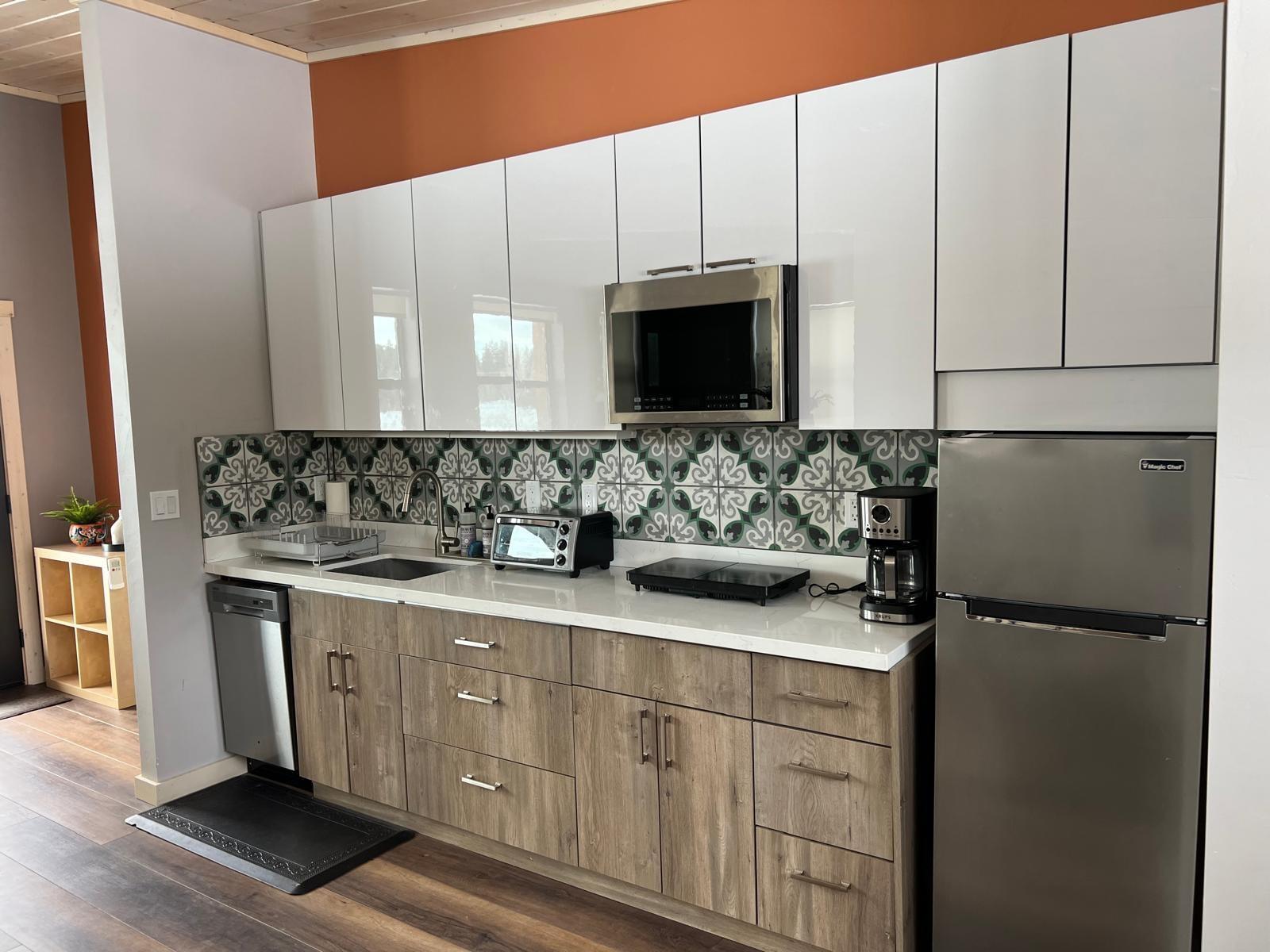 The apartment kitchen: a single galley run under gloss white cabinets, with a green and white patterned tile backsplash, dishwasher, sink, induction cooktop, microwave and full-size fridge, below a terracotta wall and a pine ceiling