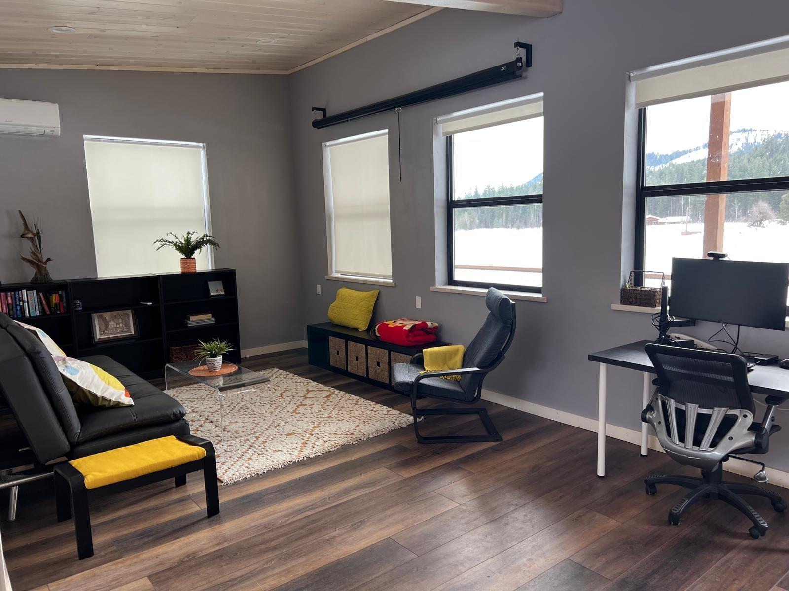 The apartment living room: a black futon and ottoman on a woven rug, bookshelves and a cushioned window seat along one wall, and a desk with a large monitor under three windows looking out over the snow to the ridge
