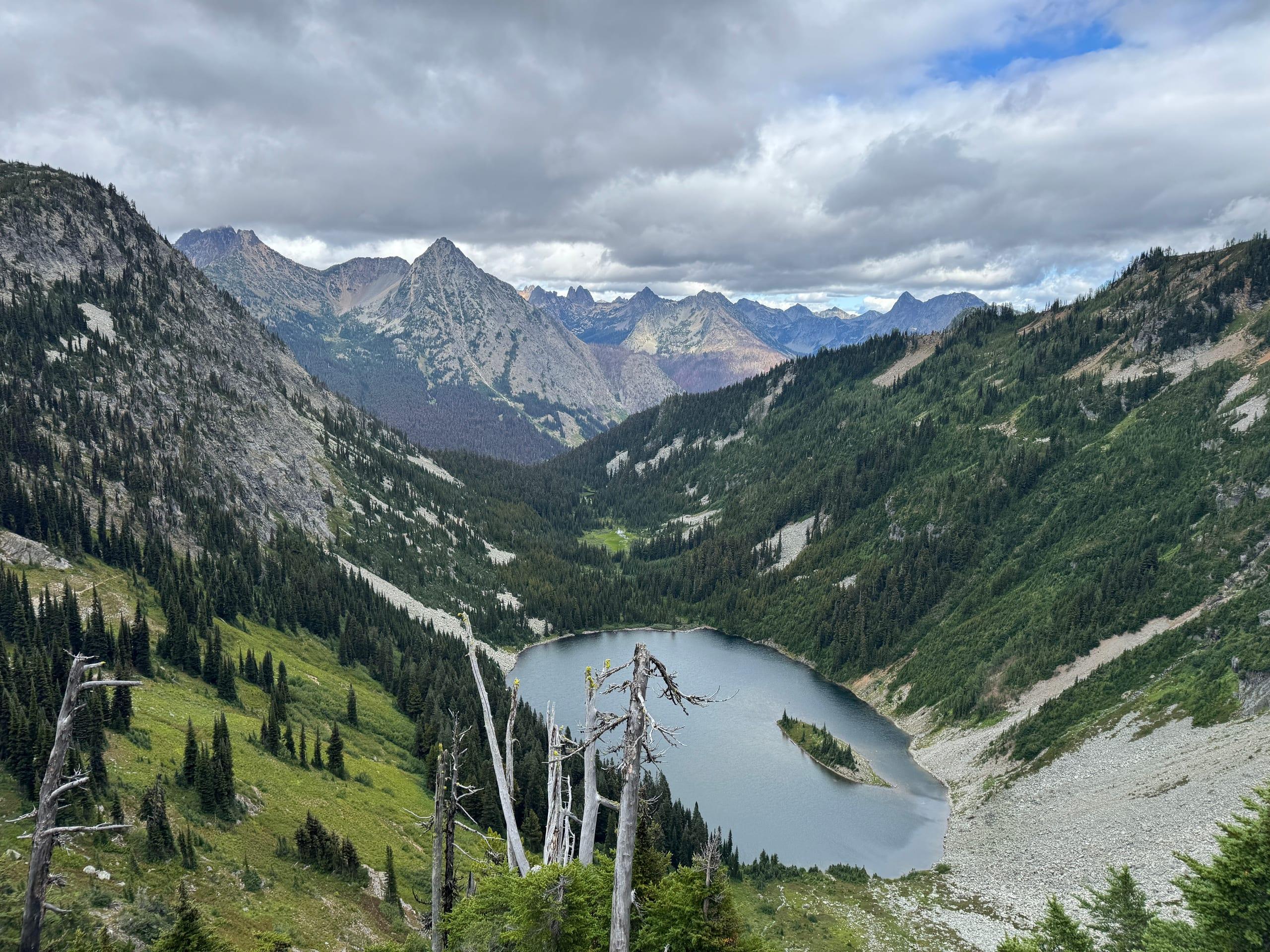 An alpine lake with a small wooded island at the head of a hanging valley, ringed by steep green slopes and scree, with row on row of North Cascades peaks beyond