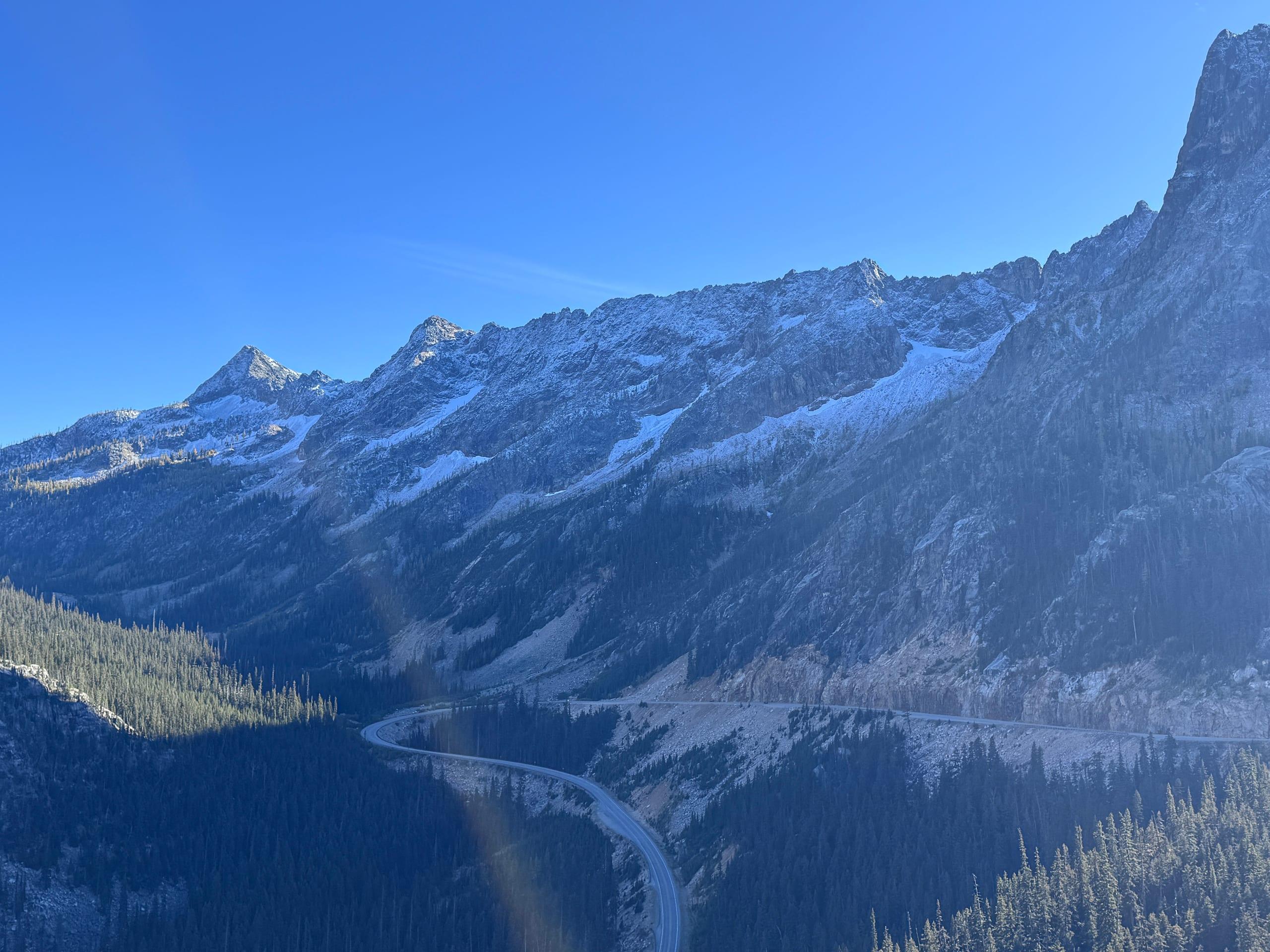A highway switchbacking far below a high ridge dusted with early snow, dark forest filling the valley floor under a clear sky