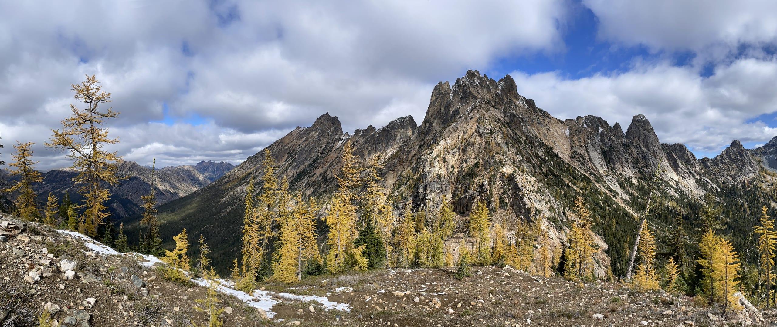 A wide view along a ridge of golden larches to a line of jagged rock spires, early snow lying in the gullies and cloud breaking over the range behind