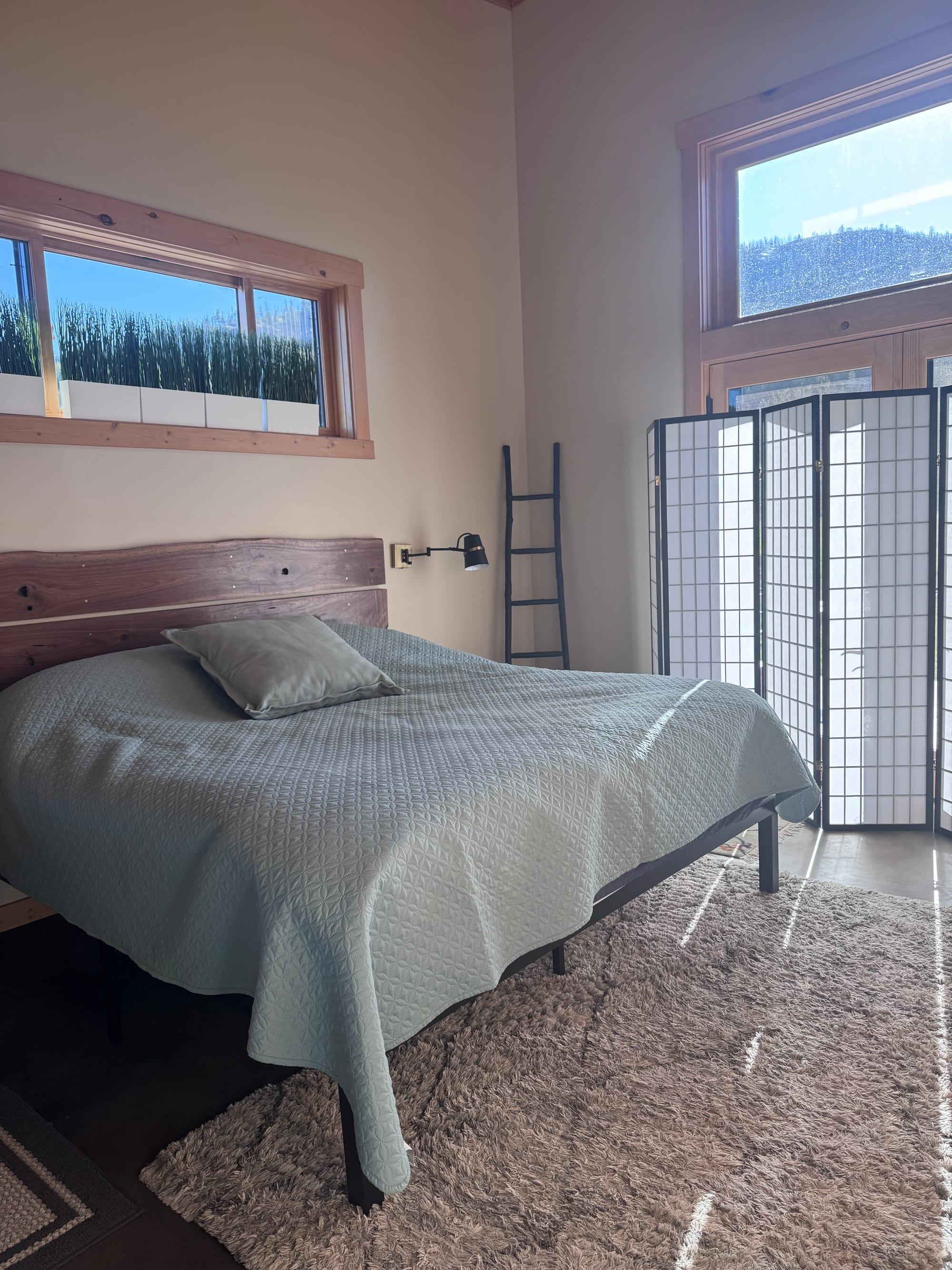 The master bedroom: a live-edge timber headboard and sage quilt, with clerestory windows above and a shoji screen onto the valley