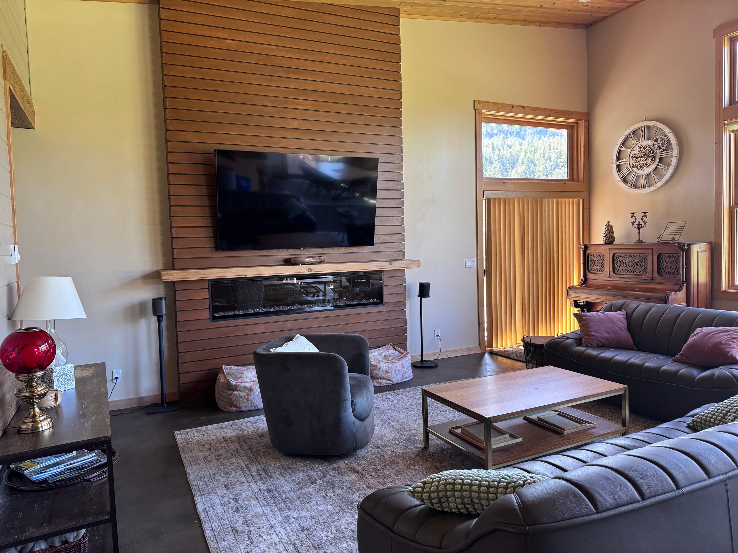 The fireplace wall: horizontal cedar slats rising to the pine ceiling around a long linear fireplace under a floating timber mantel, with a swivel armchair and leather sofas on a patterned rug