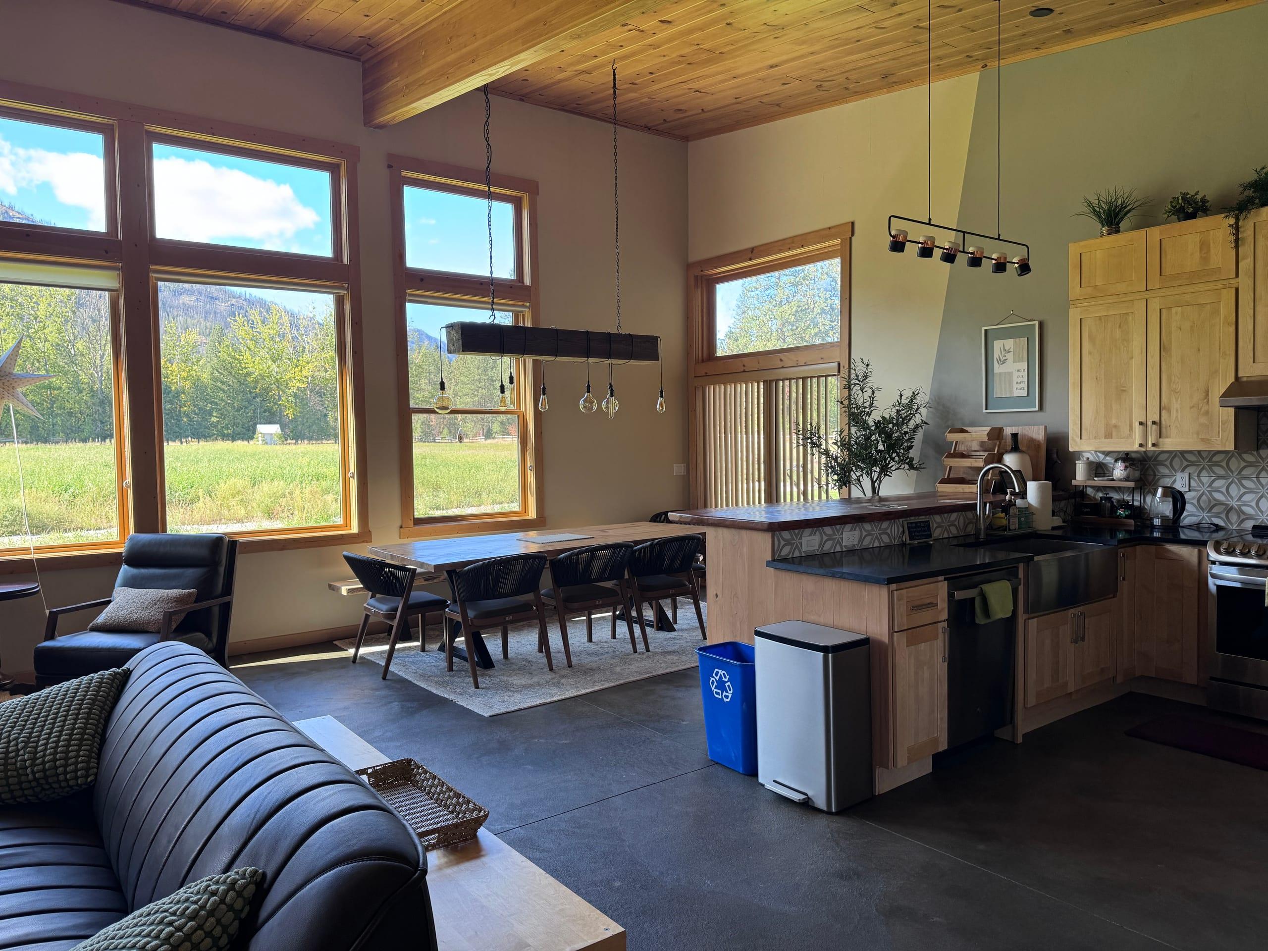 The open plan from the living area: a live-edge dining table under a beam pendant light by the meadow windows, running into the kitchen with maple cabinets, dark counters and a farmhouse sink