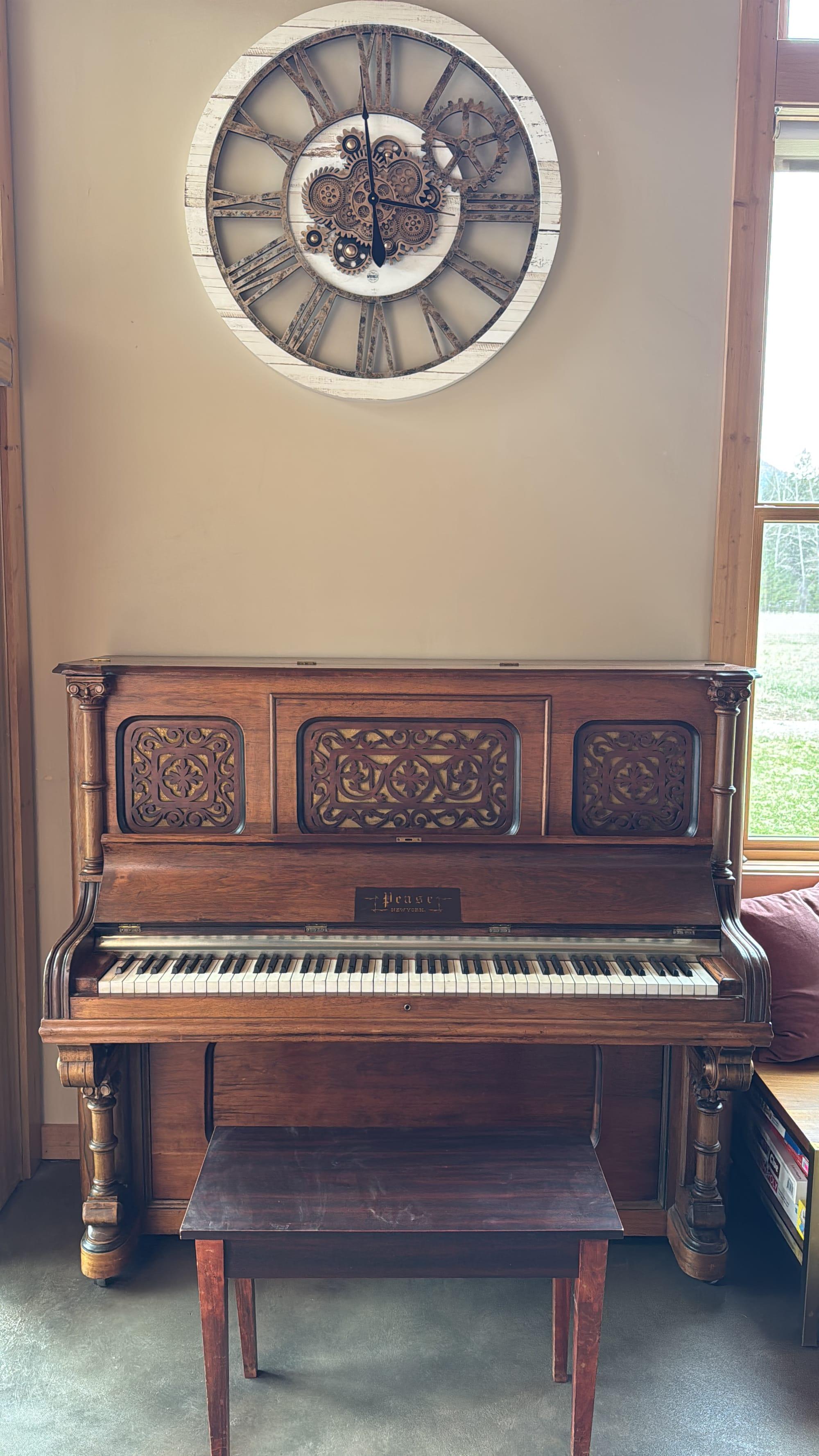 An antique carved upright piano in the great room, below a station clock, beside a window onto the meadow