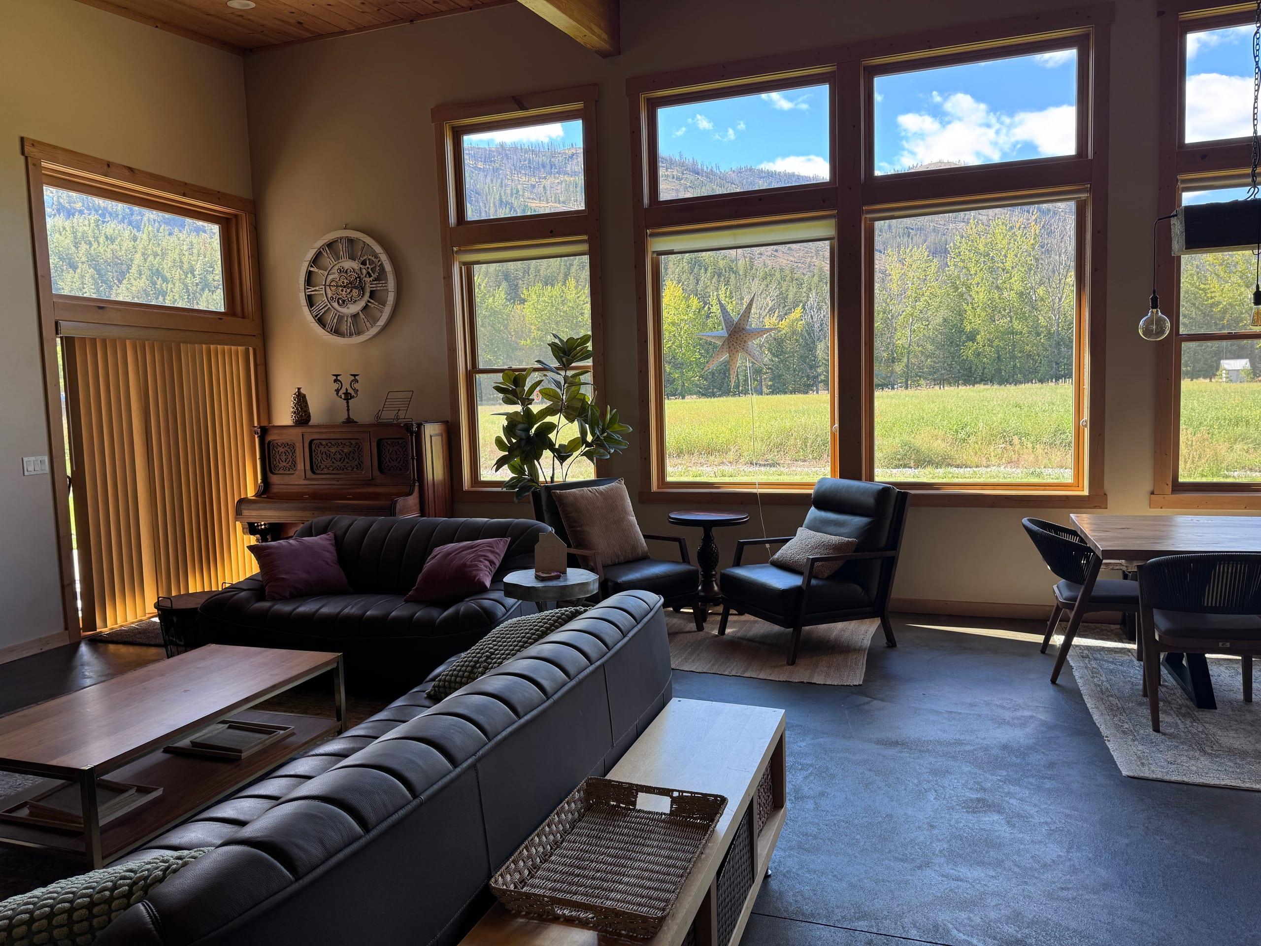 The great room in afternoon sun: two dark leather sofas and a pair of armchairs facing a wall of tall timber-framed windows onto the green meadow and the forested hillside beyond, with a carved antique piano and a skeleton wall clock to one side